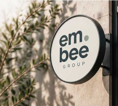 em.bee Group lettering on a round wall sign beside greenery